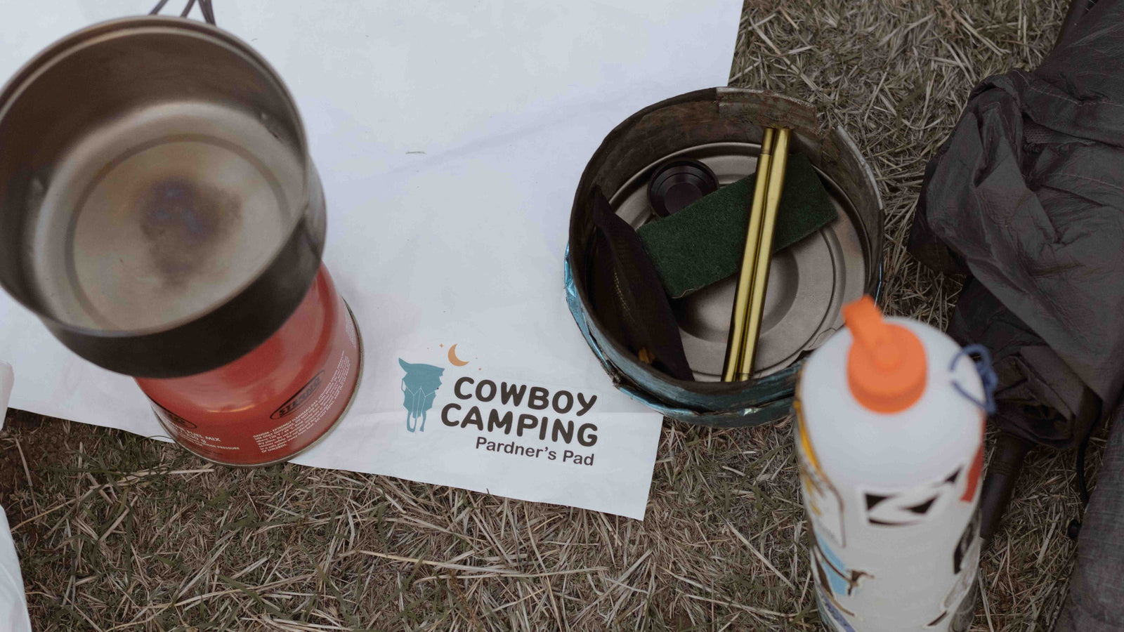 The corner of the Pardner's Pad lays on the ground surrounded by backpacking cooking tools and a ThruBottle