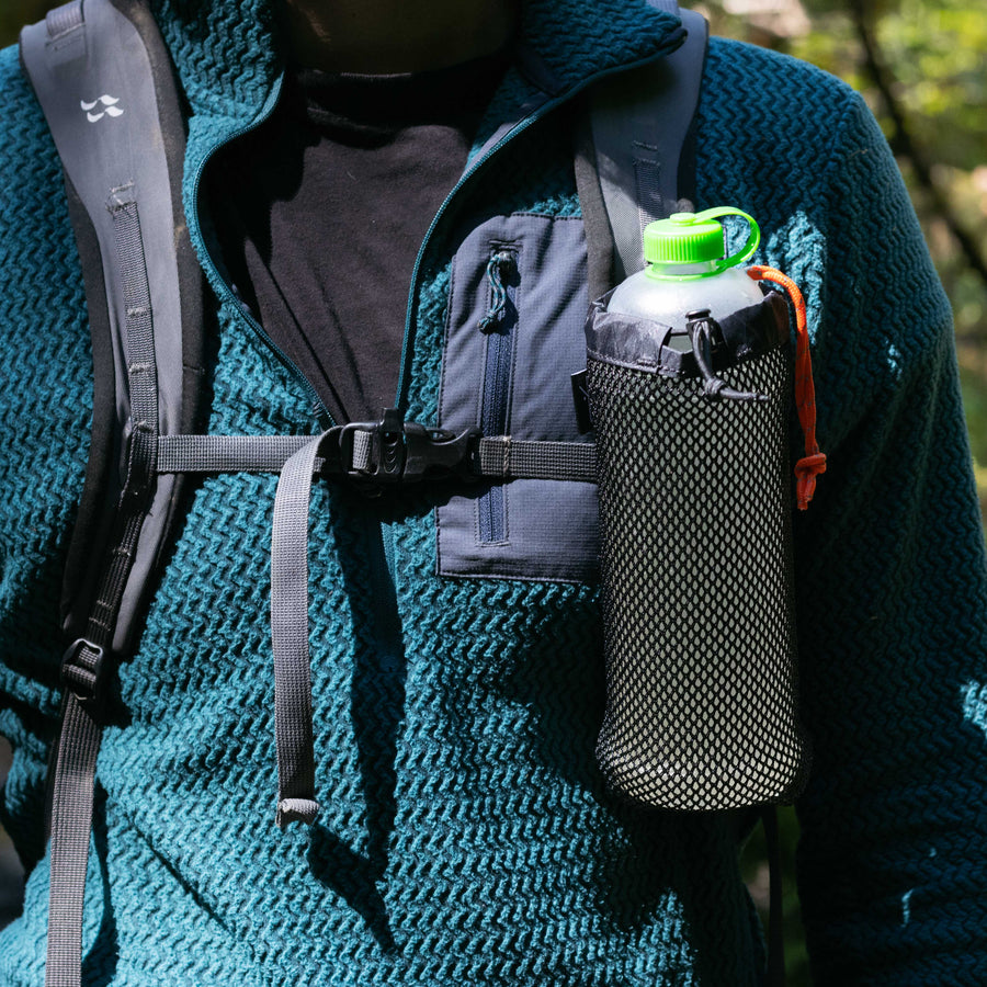 A close-up of a hiker wearing a backpack with a shoulder sleeve pocket that holds a 700ml Vesica bottle